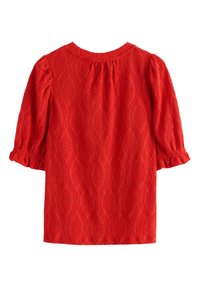 Rød tekstureret bluse med diamantformede udskæringsmønstre, rund halsudskæring og trekvartlange pufærmer med rynkede manchetter.