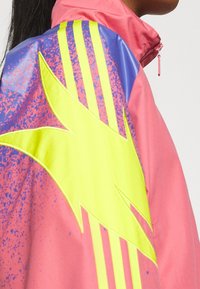 Vindjacka i rosa med en stor gul stjärndesign och blå detaljer. Har randiga detaljer på ärmarna och en dragkedja vid kragen.