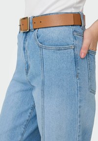 Světlé modré džíny s vysokým pasem, výrazným šitím a dvěma předními kapsami. Spárováno s opaskem z hnědé kůže se zlatou přezkou.