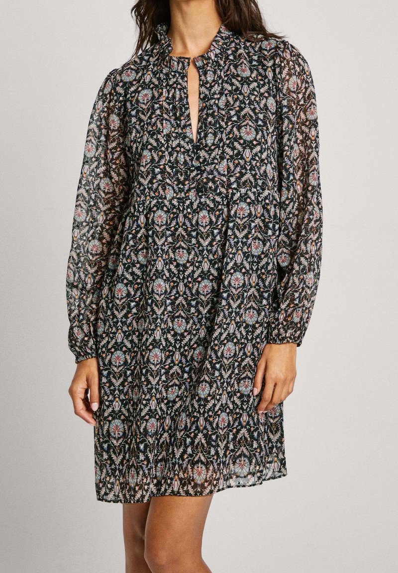 Robe noire longue à manches longues en tissu transparent avec motif floral, encolure froncée et ouverture frontale en forme de goutte, portée par une personne debout.