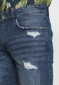 Jeans de mezclilla azul oscuro con un ajuste slim, que presentan parches desgastados en los muslos y una textura descolorida. Incluyen un bolsillo frontal y cierre de botón.