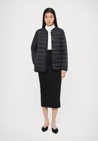 Μαύρο μπουφάν τύπου puffer με στρογγυλή λαιμόκοψη, κουμπιά με πίεση και οριζόντια ραφή, συνδυασμένο με μαύρη midi φούστα και μαύρα μυτερά παπούτσια.
