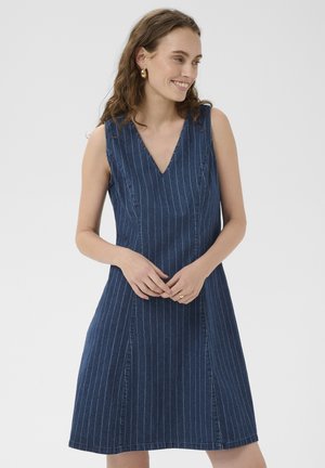 Kvinde iført ærmeløs marineblå kjole med nålestriber og V-hals, smilende med hænder løst foldet foran sig, mod en ensfarvet lys baggrund.