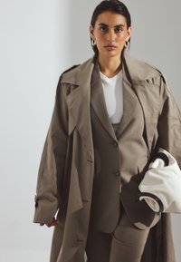 Bežový oversize trenčkot s ostrými klopami cez priliehavý taupe oblek, doplnený bielou taškou s čiernymi akcentmi.