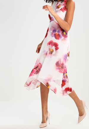 Frau trägt ein weißes ärmelloses Kleid mit großen rosa und violetten Blumenmustern, geht und trägt beige High Heels.