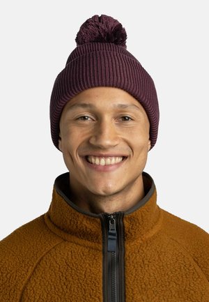 Gorro de punto burdeos con un pompón en la parte superior, usado por una persona con una chaqueta de forro polar naranja con cierre de cremallera y cuello alto.