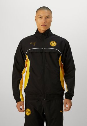 Black and yellow track jacket with a zip front, featuring a high collar, side stripes, and logos on the chest and pants. Lightweight fabric.