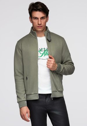 Mężczyzna w jasnzie zielonej kurtce na zamek, nałożonej na białą koszulkę z nadrukiem, oraz czarnych spodniach, stojący na jednolitym tle.