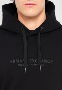 Homme portant un sweat à capuche noir avec cordons, avec le texte « ARMANI EXCHANGE MILANO / NEW YORK » sur la poitrine, cadré sous le nez.