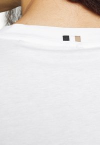 T-shirt bianca in cotone con colletto girocollo e un piccolo cartellino con tre strisce orizzontali: nera, bianca e beige. Tessuto semplice e liscio.
