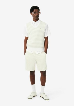 Mann mit übergroßen Sonnenbrillen, weißem kurzärmligem Hemd, cremefarbener Strickweste, cremefarbenen Shorts, weißen Socken und weißen Turnschuhen, stehend mit den Händen in den Taschen.