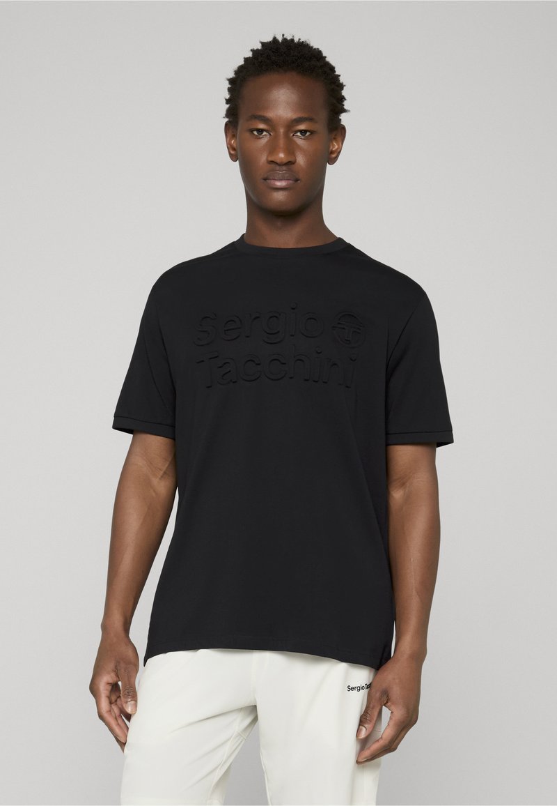 Jeune homme portant un t-shirt noir Sergio Tacchini et un pantalon blanc, debout devant un fond gris uni.