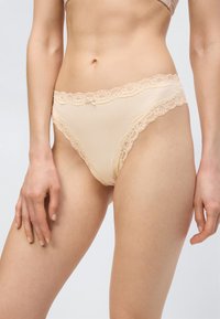 Γυναίκα που φοράει μπεζ εσώρουχα με δαντελένια διακόσμηση, στέκεται με τα χέρια χαλαρά δίπλα στο σώμα της μπροστά σε απλό φόντο.