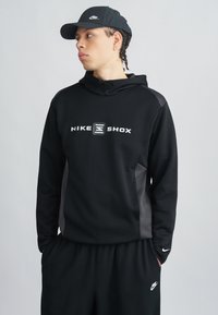Μαύρο φούτερ Nike με το λογότυπο "NIKE SHOX" σε λευκό, με φερμουάρ στο γιακά, πλαϊνές επενδύσεις και μαύρο παντελόνι φόρμας που ταιριάζει.