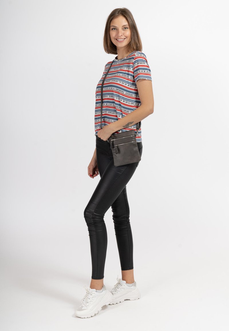 Gestreept T-shirt in verschillende kleuren, gecombineerd met glanzende zwarte leggings en witte sneakers. Model houdt een kleine, grijze schoudertas vast.