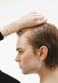 Pendientes de aro de plata con un acento de estrella, usados junto a pulseras de cadena de plata que cuentan con un crucifijo y un charm circular. Textura suave, diseño minimalista.