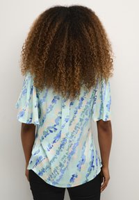 Blusa azzurra con motivi floreali in diverse tonalità di blu e rosa. Presenta maniche corte a volant e un'orlo arrotondato.