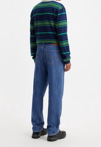 Námořnické pruhované tričko s dlouhým rukávem v zelenohnědých a oranžových pruzích, spárované s modrými denimovými džíny s volným střihem. Celkový vzhled dotvářejí černé tenisky.