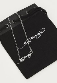 Ασημένια αλυσίδα με κρεμαστό στυλιζαρισμένο «Ed Hardy». Η αλυσίδα βρίσκεται πάνω σε μία μαύρη σακούλα από ύφασμα με λευκή επωνυμία και κορδόνι.