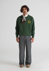 Zielony sweter z okrągłym dekoltem i żółtym haftem, w połączeniu z szarymi spodniami w paski i wzorzystą chustą związaną wokół szyi.