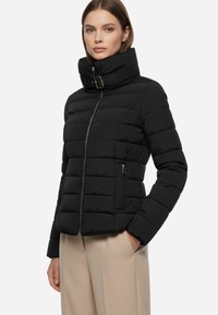 Giubbotto nero imbottito con colletto alto, texture a cuciture orizzontali, zip frontale e tasche laterali. Presenta un dettaglio a fibbia sul colletto.