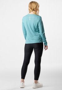 Top atletico a maniche lunghe di colore azzurro chiaro, realizzato in un materiale liscio, abbinato a leggings neri aderenti. Accenti bianchi e logo visibili sull'abbigliamento.