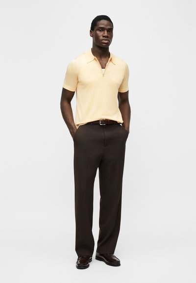 Άνδρας που στέκεται με τα χέρια στις τσέπες, φορώντας ανοιχτό κίτρινο κοντομάνικο πουκάμισο με γιακά, σκούρο καφέ παντελόνι και σκούρα καφέ παπούτσια.