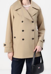 Beige trenchcoat med dubbelknäppt design, svarta knappar, bred krage och sidfickor. Bärs med mörka jeans och en svart väska.