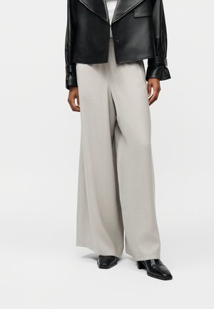 Person trägt helle beige Hosen mit weitem Bein, schwarze hochhackige Schuhe mit quadratischer Spitze und eine schwarze Lederjacke mit gerafften Ärmeln.