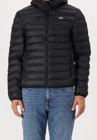 Černá prošívaná bunda s kapucí, předním zipem, bočními kapsami a zeleným logem na hrudi. Hladký, lesklý povrch.