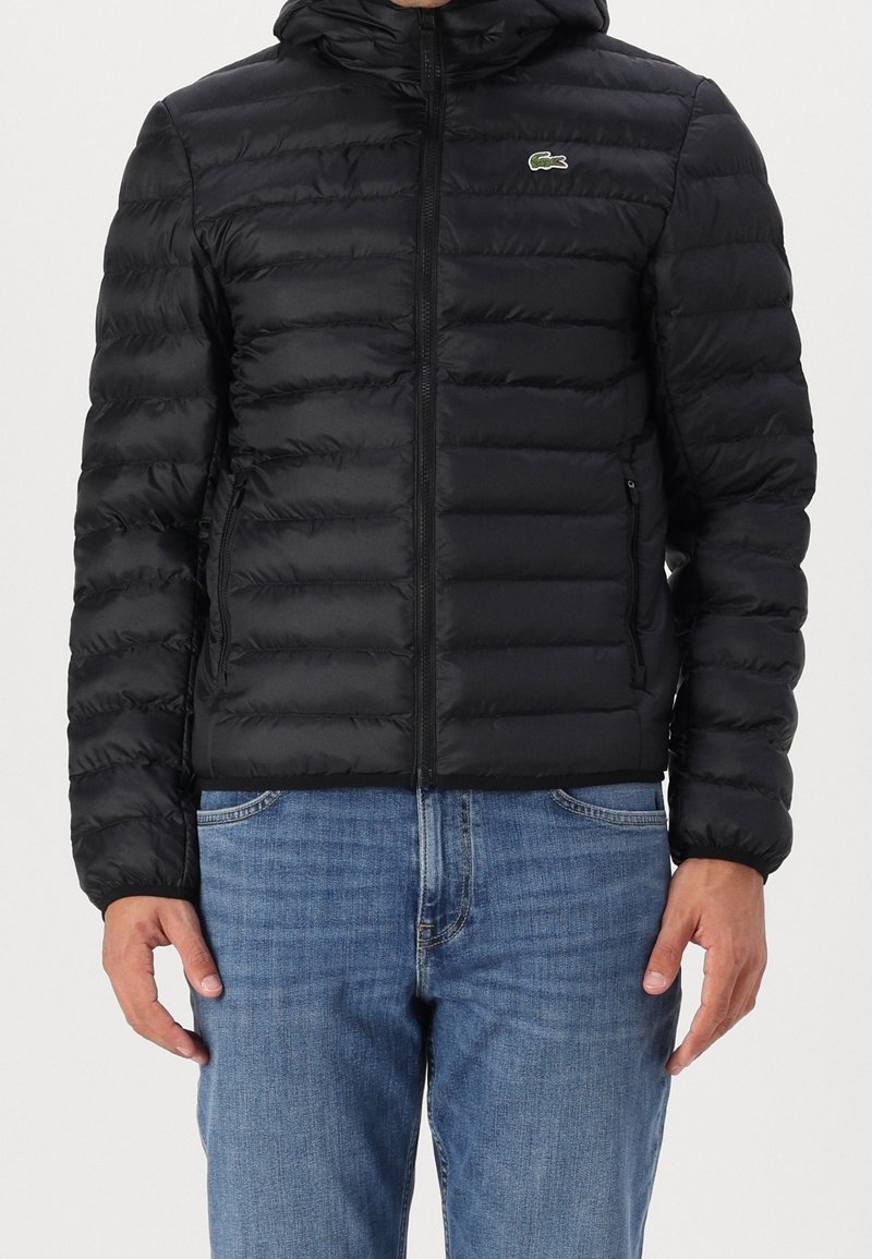 Černá prošívaná bunda s kapucí, předním zipem, bočními kapsami a zeleným logem na hrudi. Hladký, lesklý povrch.