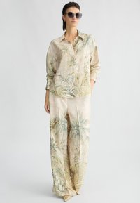 Blouse légère à motifs et pantalon large en beige doux avec des imprimés botaniques. Les accessoires comprennent des lunettes de soleil sombres et des chaussures à bout pointu.