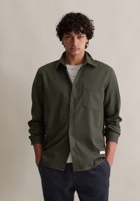 Giovane uomo con capelli ricci che indossa una camicia verde scuro a bottoni, una maglietta bianca sotto e pantaloni scuri a coste, in piedi con le mani in tasca.