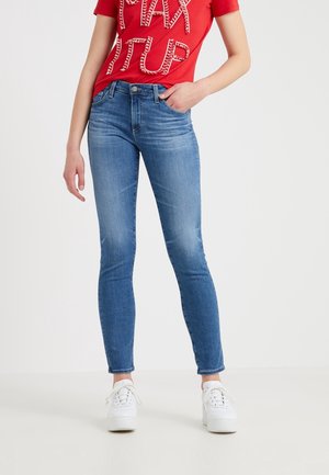 Kobieta w dopasowanych niebieskich dżinsach, czerwonej koszulce z napisem "PODNIEŚ PODATKI" i białych trampkach, stoi z jedną ręką w kieszeni.