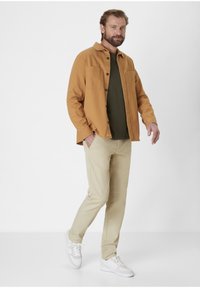 Khaki trousers, olive green shirt, and a mustard yellow button-up jacket. White sneakers with textured soles. Neutral background.