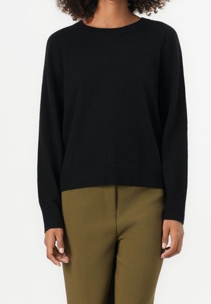 Osoba ubrana w czarny sweter z długim rękawem i okrągłym dekoltem oraz oliwkowo-zielone spodnie, stojąca na białym tle.