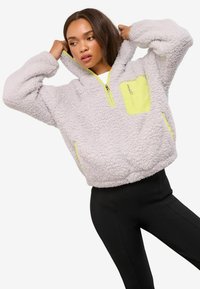 Heledhalli fliisist pullover, millel on osaline tõmblemine, neoonkollased aktsendid krael ja taskul, omab pehmet, tekstuurset pind.