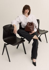 Braune Leder-Eimer-Tasche mit Zugband, getragen mit einer weißen Bluse mit Rüschenkragen, schwarzen weit geschnittenen Hosen und spitzen schwarzen Pumps.