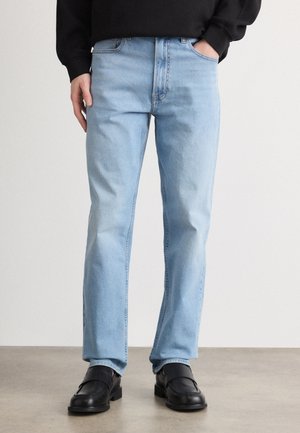 Šviesiai mėlyni džinsai su tiesiu kirpimu, turintys penkis kišenes, užtrauktuką ir subtilų blukimą. Dėvimi su juodais neslidaus pado bateliais.