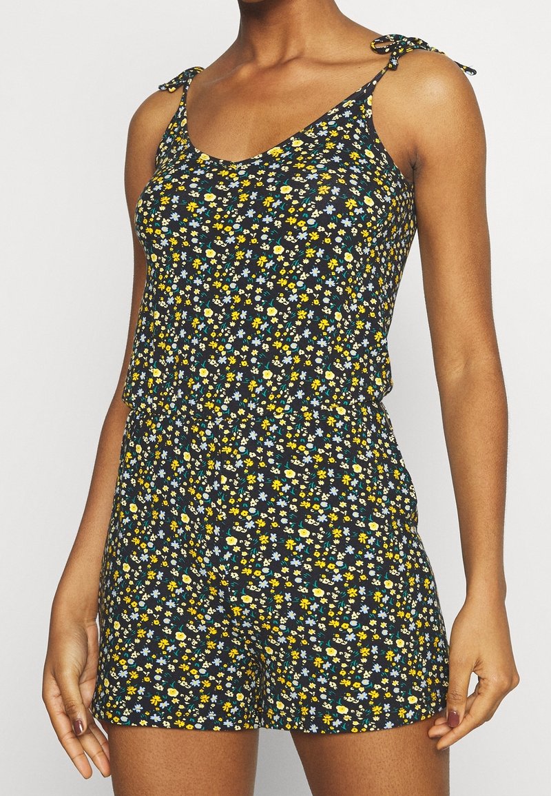 Femme portant une combinaison-short noire à motifs floraux avec de petites fleurs jaunes, bleues et blanches et des bretelles fines.