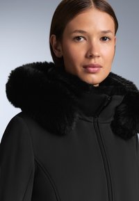 Crni kaput s plišanim lažnim krznenim kapuljačom. Glatka tkanina, prednje zatvaranje na zip, i strukturirani siluet. Mekana tekstura i precizno dizajnirani detalji.