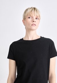 Melna kokvilnas t-krekls ar apaļu kakla izgriezumu un īsām piedurknēm, ar izkliedētām pērļu detaļām, kas piešķir tekstūru. Minimalistisks dizains.