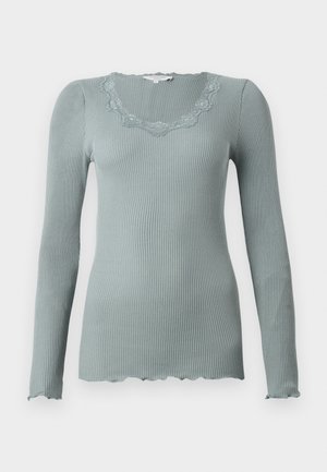 Langermet, ribbestrikket topp i dempet grønn med en bølget, blondebesatt halslinning. Myk tekstur og tettsittende design.