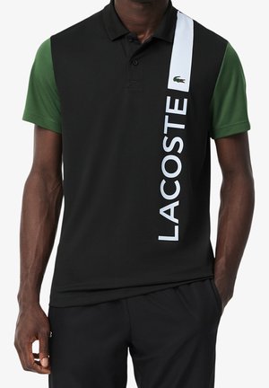 Person trägt ein schwarzes Polo-Shirt mit grünen Ärmeln und weißem vertikalem "LACOSTE" Text und Logo auf der rechten Vorderseite.