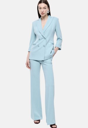 Donna che indossa un blazer doppiopetto azzurro chiaro con pantaloni a gamba larga abbinati e grandi orecchini rotondi, in piedi con una mano in tasca.