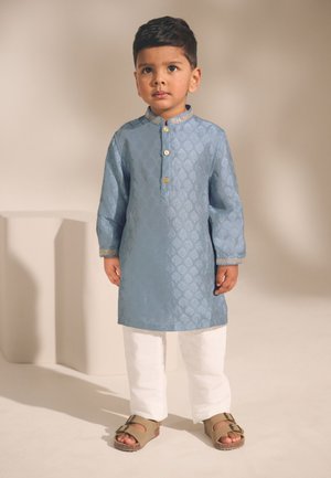 Giovane ragazzo in piedi, che indossa una kurta azzurro chiaro con motivi, bottoni dorati, pantaloni bianchi e sandali marroni su uno sfondo semplice.