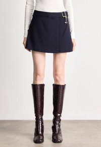 Σαλοπέτα ναυτικού χρώματος με μινιμαλιστικό σχέδιο, διαθέτει ζώνη με χρυσό κούμπωμα και με γοητεία σε σχήμα αστεριού. Συνδυάζεται με λαμπερά, σκούρα μπορντό μπότες μέχρι το γόνατο.