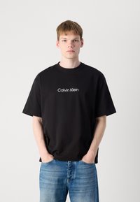 Svart bomull T-skjorte med rund hals og korte ermer, med en hvit "Calvin Klein" logo sentrert på brystet. Båret sammen med blå denimjeans.