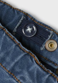 Denim tkanina v temno modri barvi z vidnim gumbom in zaponko v zlatem odtenku. Oranžni šivi ob robu dodajajo kontrast.