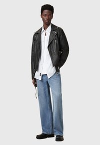 Zwart leren motorjack over een wit overhemd met knopen, gecombineerd met lichtblauwe wijde jeans en zwarte schoenen.